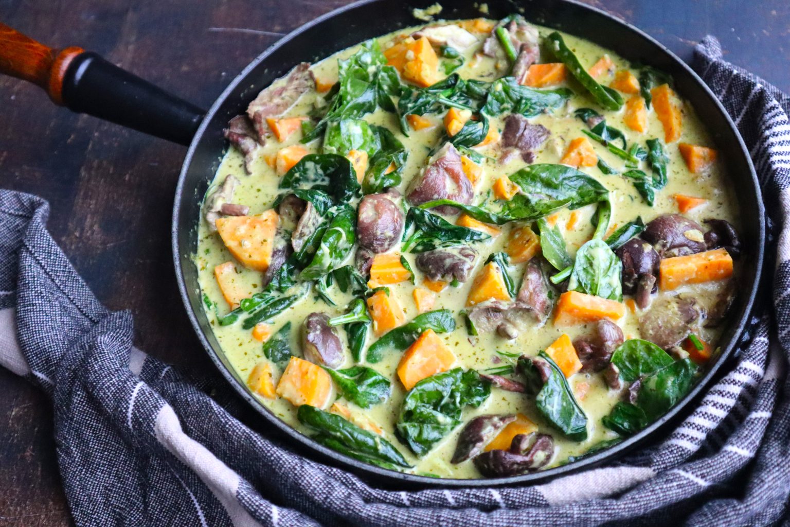 A skillet filled with a creamy stew containing chunks of sweet potato, spinach leaves, and pieces of meat, resting on a dark wooden surface beside a striped cloth.