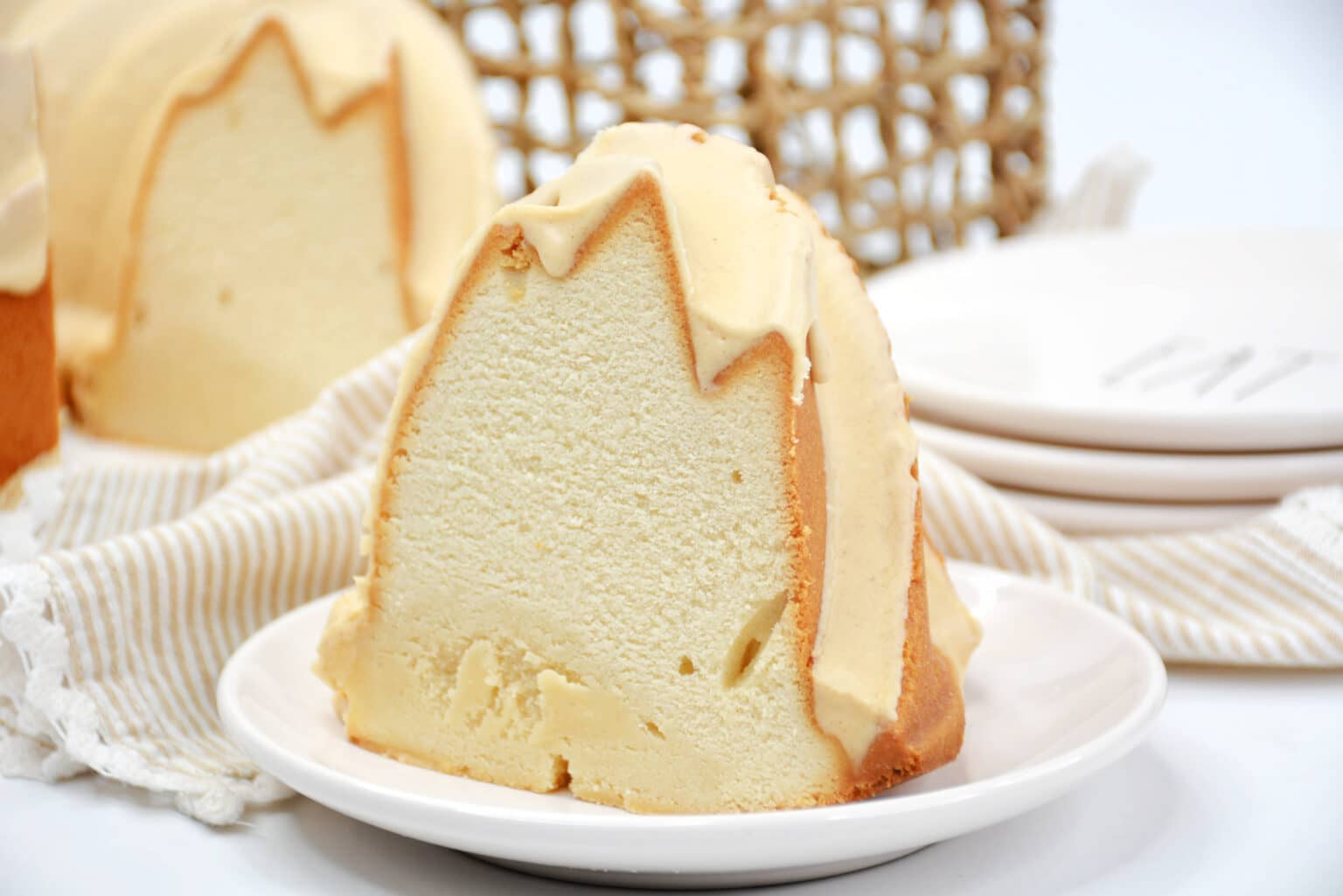 A slice of bundt cake with light-colored icing sits on a white plate, with more cake, plates, and a woven basket in the background.