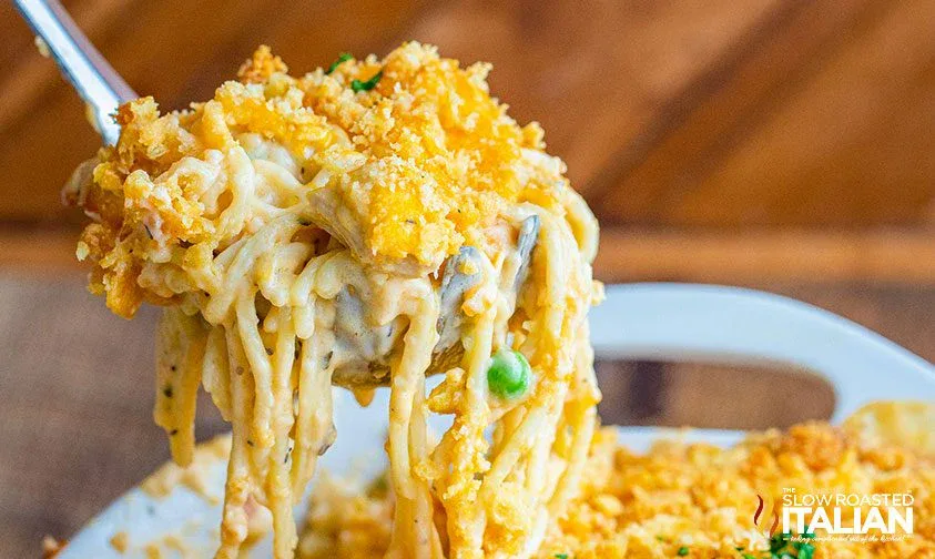 A close-up of a spoonful of creamy baked spaghetti casserole topped with a golden breadcrumb crust, with peas and pieces of chicken visible.