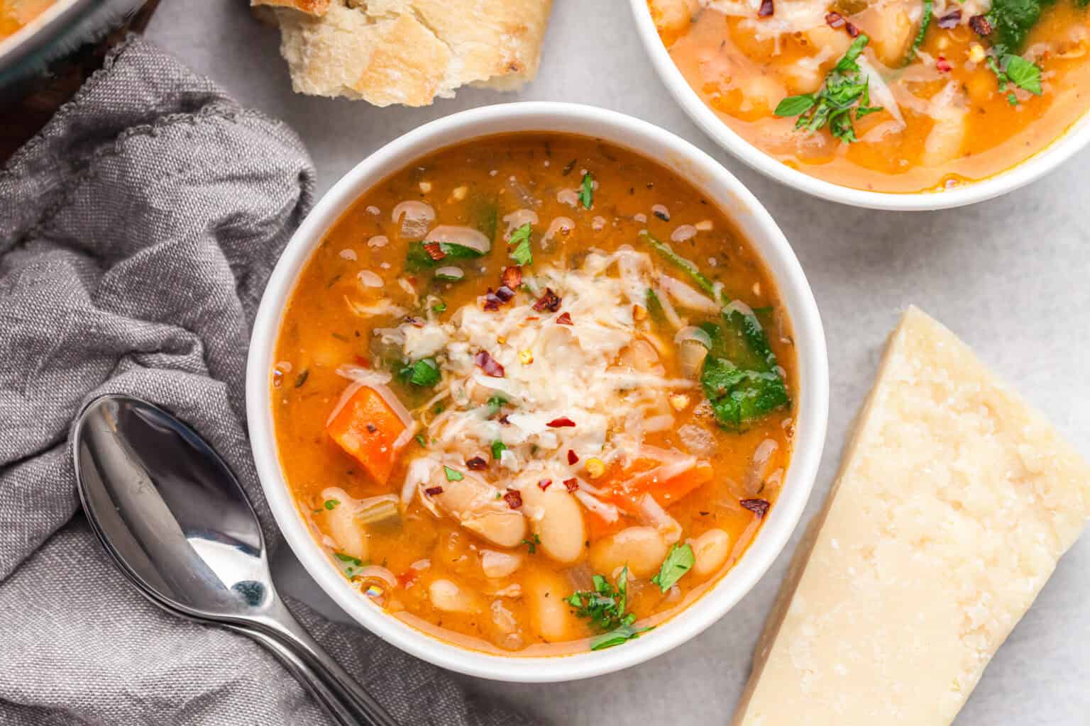 A bowl of bean and vegetable soup topped with grated cheese, surrounded by a spoon, bread, a chunk of cheese, and a gray napkin.
