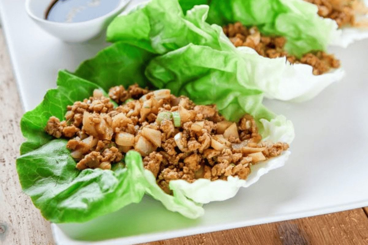 Three lettuce wraps filled with seasoned ground meat and diced vegetables are arranged on a white plate, with a small dish of sauce in the background.