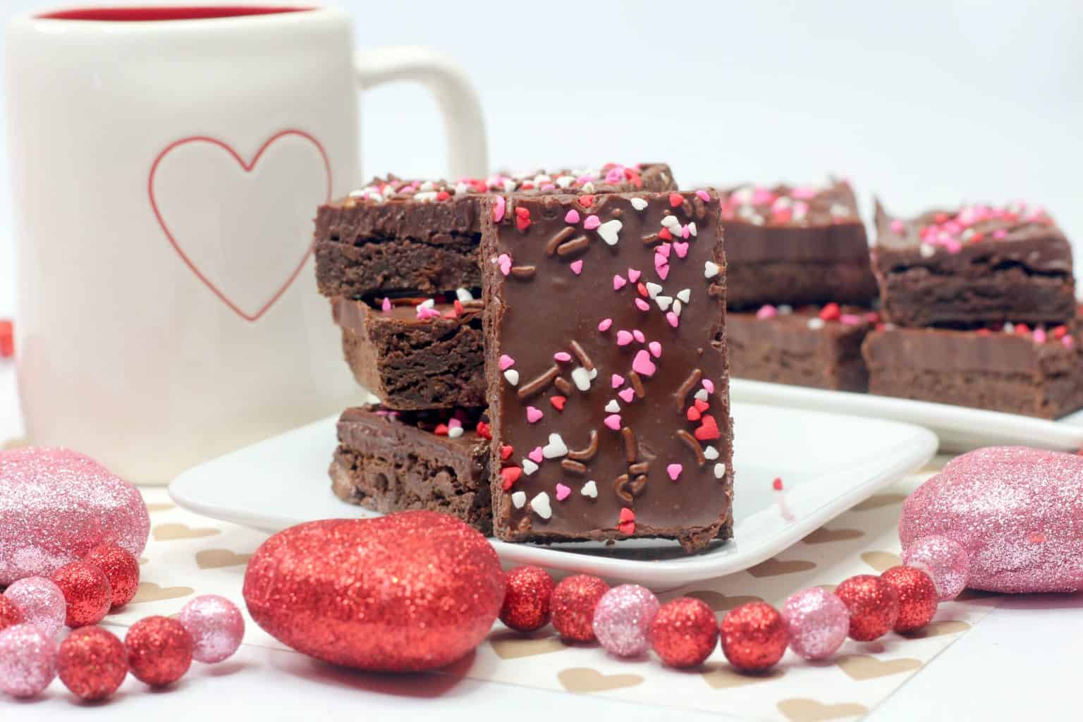 A plate of chocolate brownies topped with heart-shaped sprinkles is displayed next to a white mug with a heart design and pink glittery decorations.