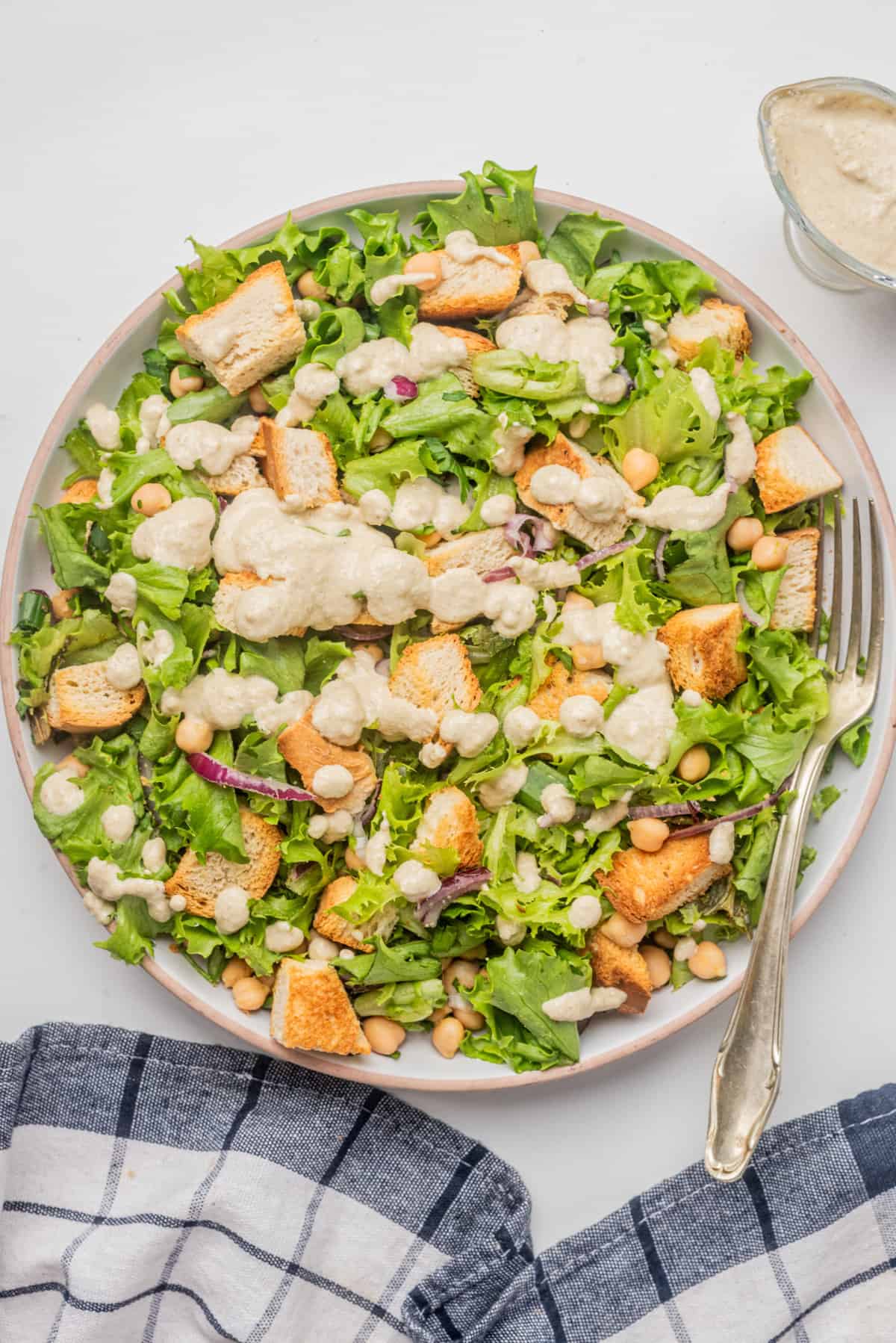 A plate of green salad with chickpeas, croutons, and a creamy dressing, served with a fork on the side and a blue checked napkin nearby.