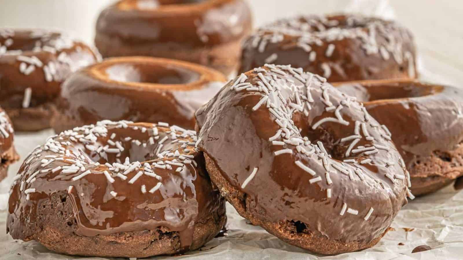 Chocolate-glazed donuts with white sprinkles are arranged on a sheet of parchment paper.