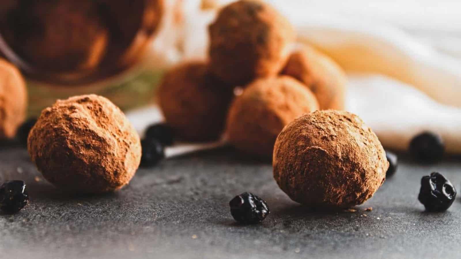 Chocolate truffles coated in cocoa powder are arranged on a dark surface with a few dried berries scattered around.