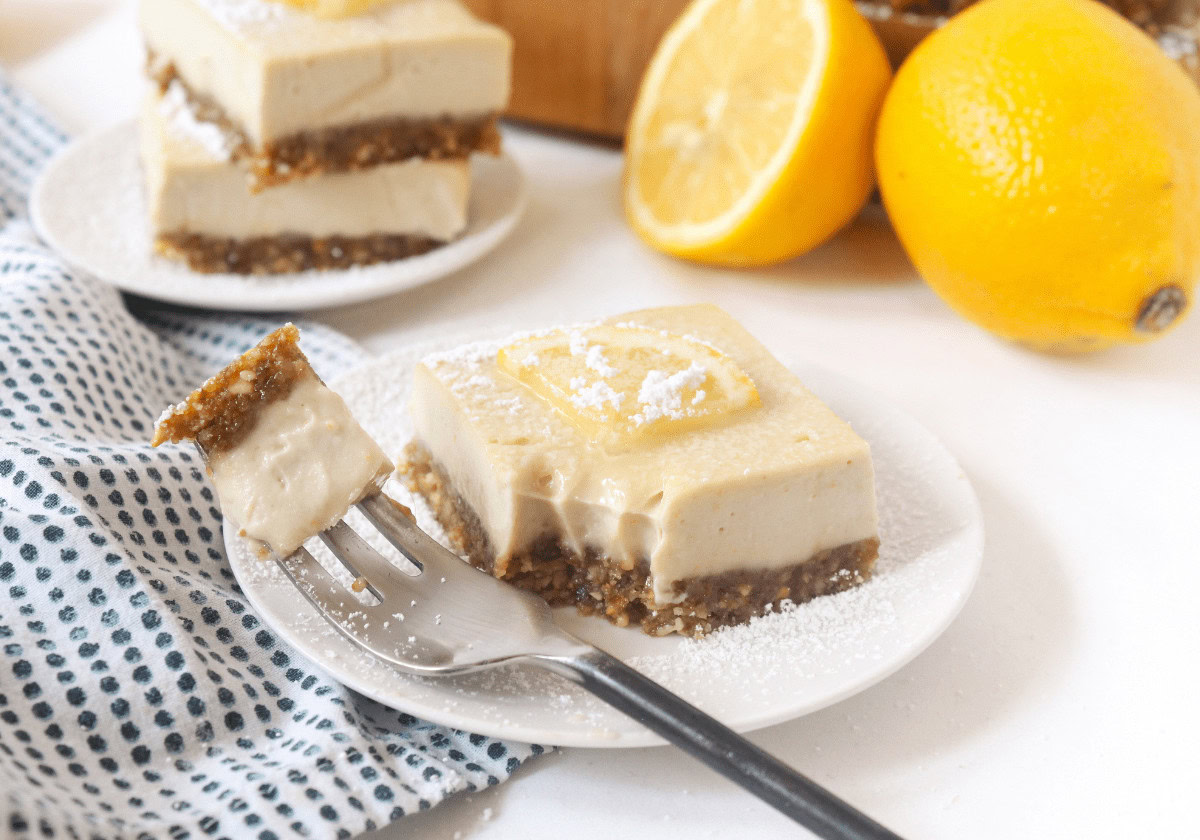 A square slice of lemon cheesecake with a date-nut crust on a white plate, garnished with a lemon slice and powdered sugar, with a fork and fresh lemons nearby.