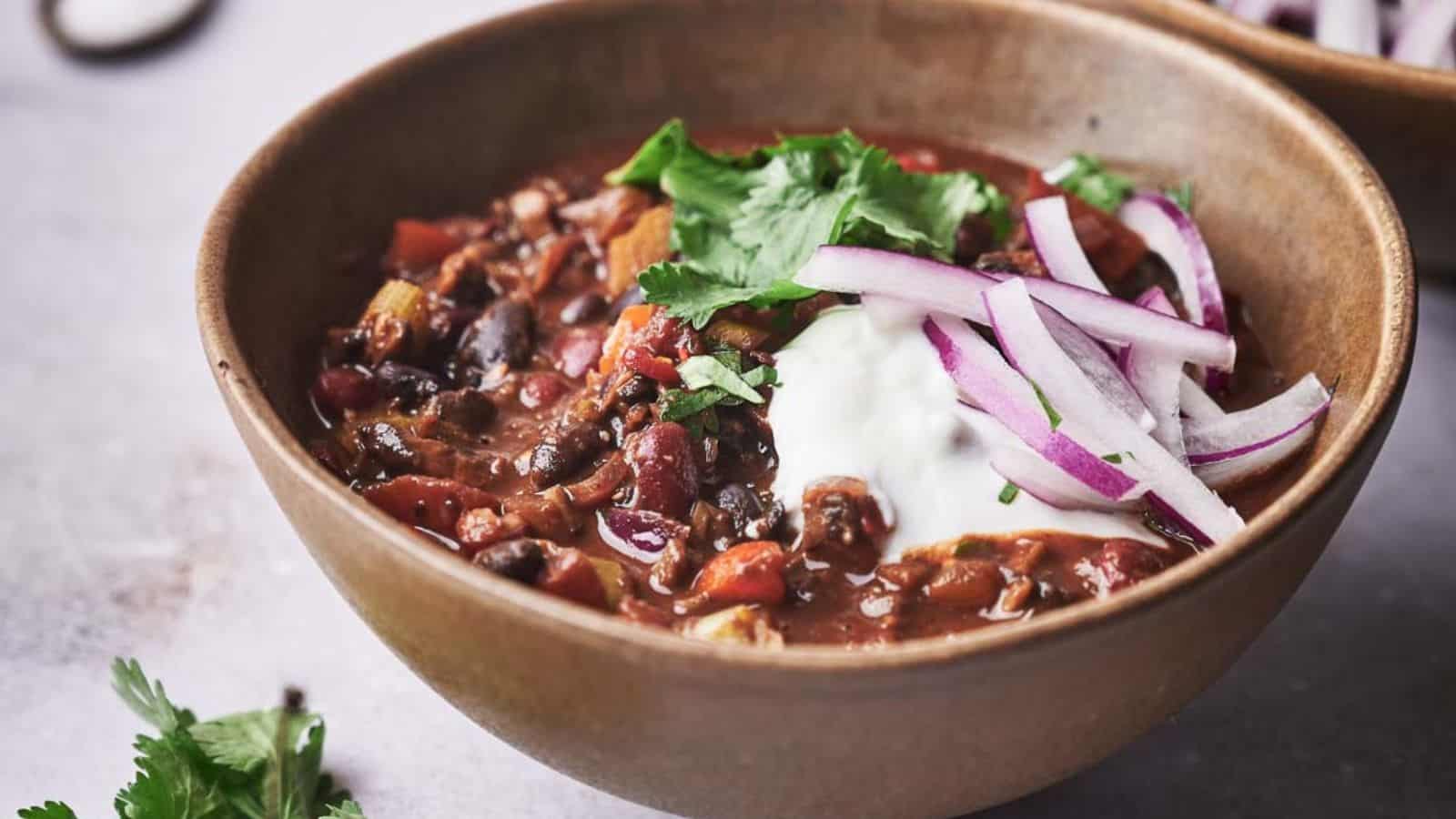 A bowl of chili topped with sour cream, sliced red onions, and fresh cilantro sits on a light surface.