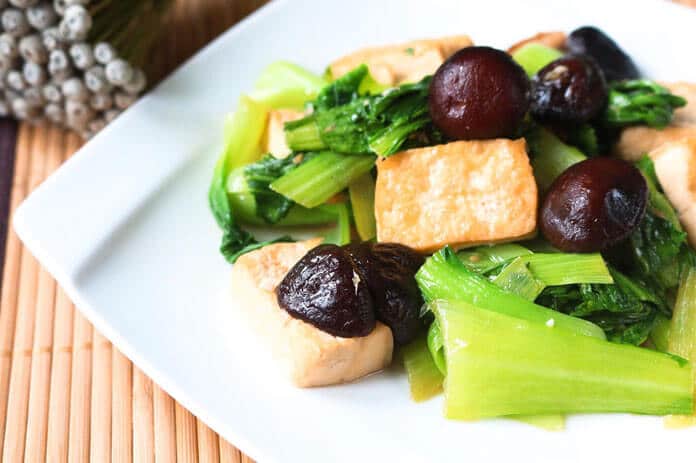 A plate of stir-fried tofu, mushrooms, and leafy greens is served on a white dish placed on a bamboo mat.