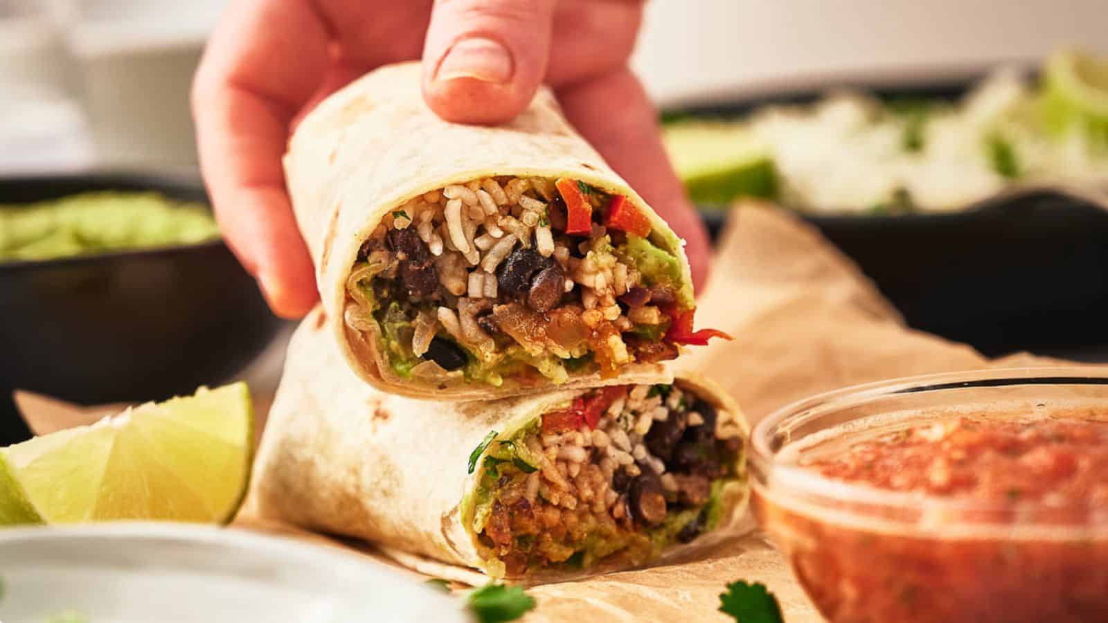 A hand holds a burrito cut in half, showing rice, black beans, vegetables, and guacamole inside; salsa and lime wedges are nearby.