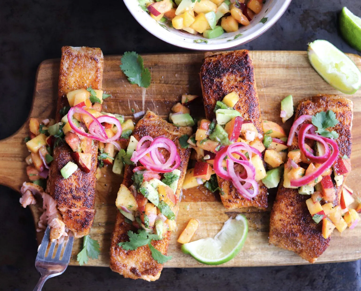Four seasoned salmon fillets on a wooden board topped with avocado-peach salsa and pickled onions, garnished with cilantro and lime wedges, with a fork and salsa bowl nearby.