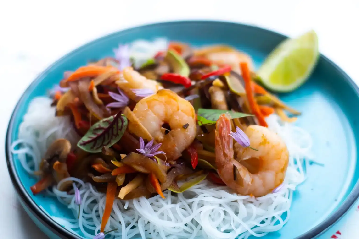A plate of rice noodles topped with shrimp, sautรฉed vegetables, edible flowers, and herbs, with a lime wedge on the side.
