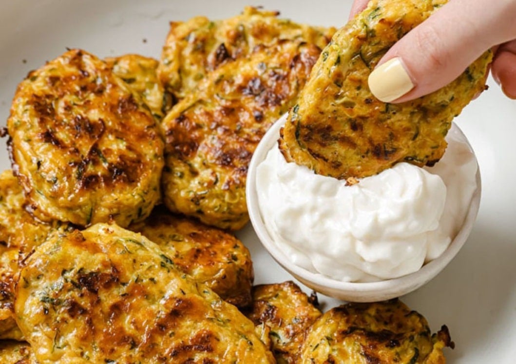 A hand dipping a zucchini fritter into a small bowl of white sauce, with more fritters on a plate in the background.