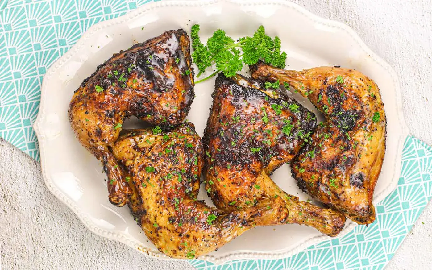 Four grilled chicken leg quarters with herbs served on a white platter, garnished with parsley, on a patterned light blue and white cloth.