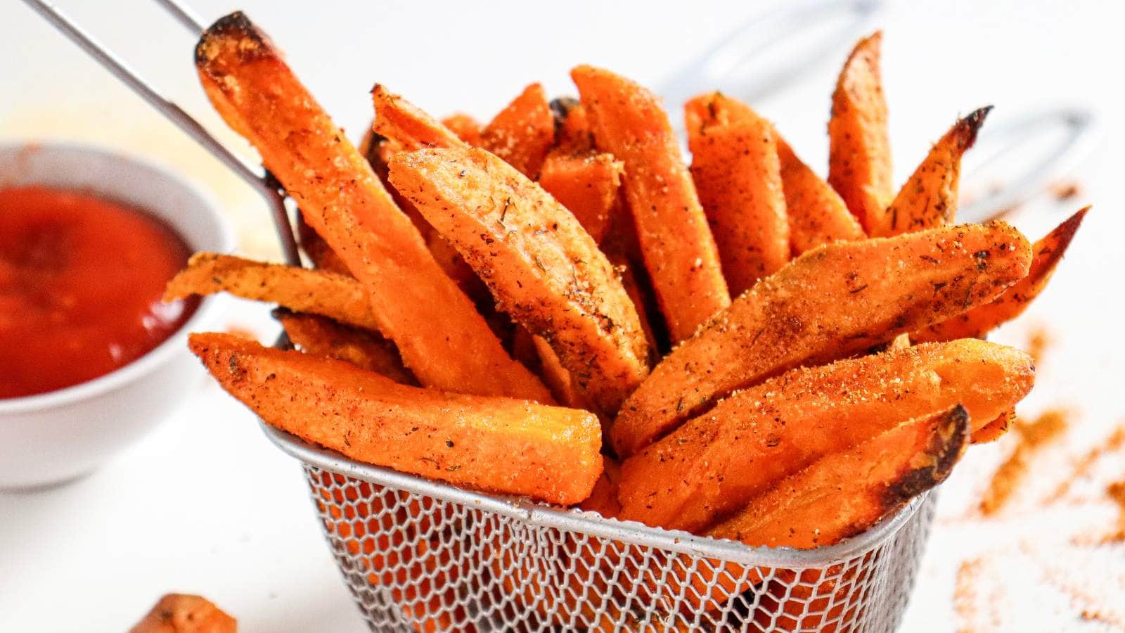 Seasoned sweet potato fries in a metal basket with a small bowl of ketchup in the background.