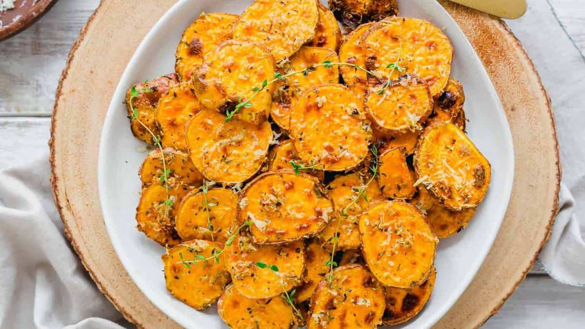 A white plate filled with roasted sweet potato rounds garnished with herbs and grated cheese, placed on a wooden charger.