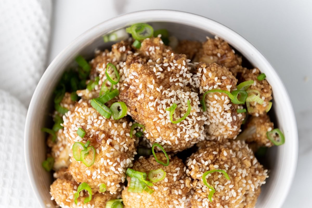 A bowl of breaded and baked cauliflower florets topped with sesame seeds and sliced green onions.