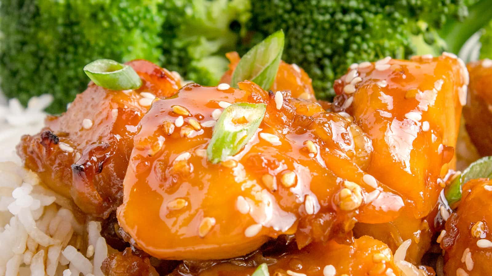 Close-up of glazed chicken pieces with sesame seeds and chopped green onions, served over rice, with broccoli in the background.