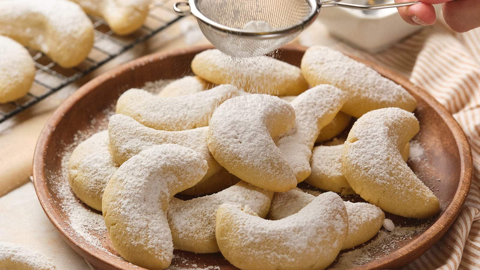 A plate of crescent-shaped cookies dusted with powdered sugar, with a hand sifting more sugar over them.