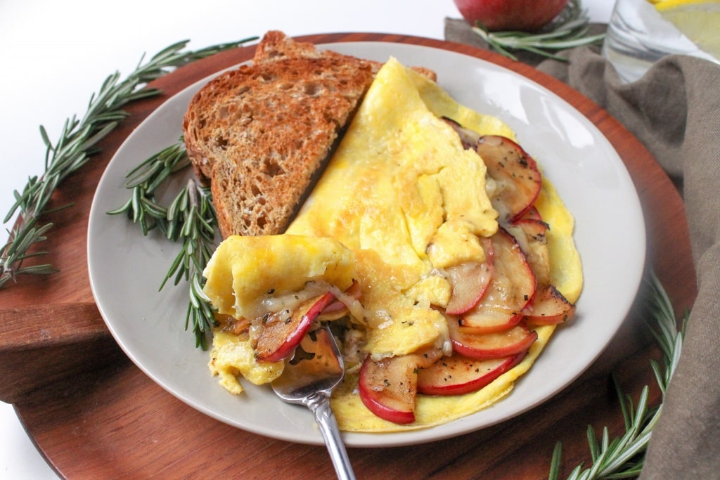 A plate with an omelette filled with sautรฉed apples and onions, served with two slices of toasted whole grain bread, garnished with fresh rosemary.