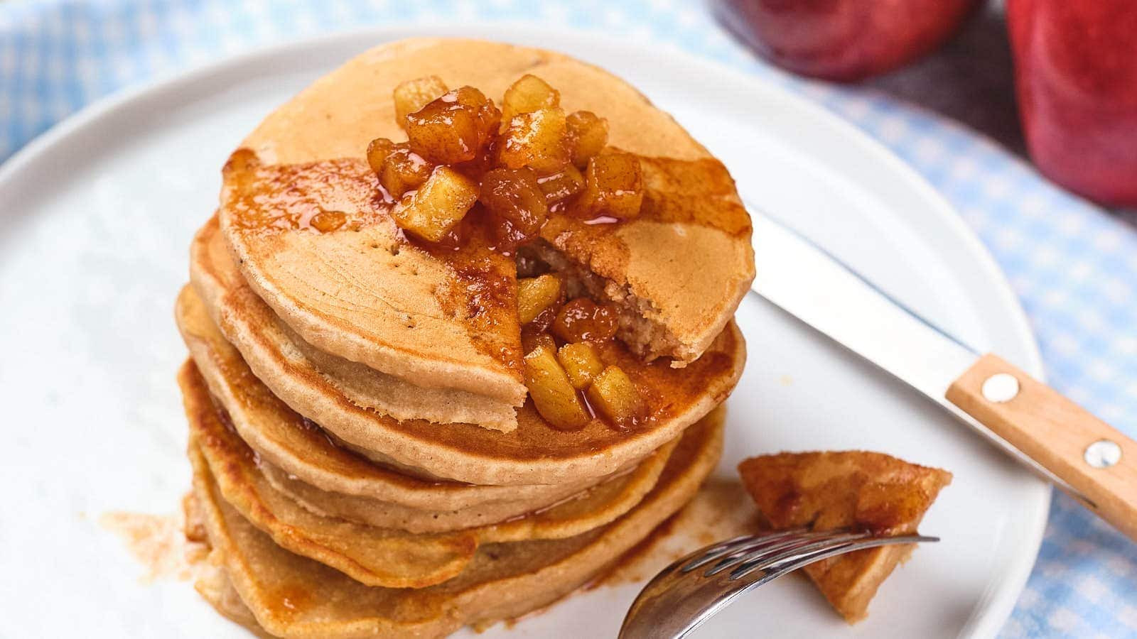A stack of pancakes topped with diced caramelized apples, with a fork holding a small piece and a knife resting on the plate.