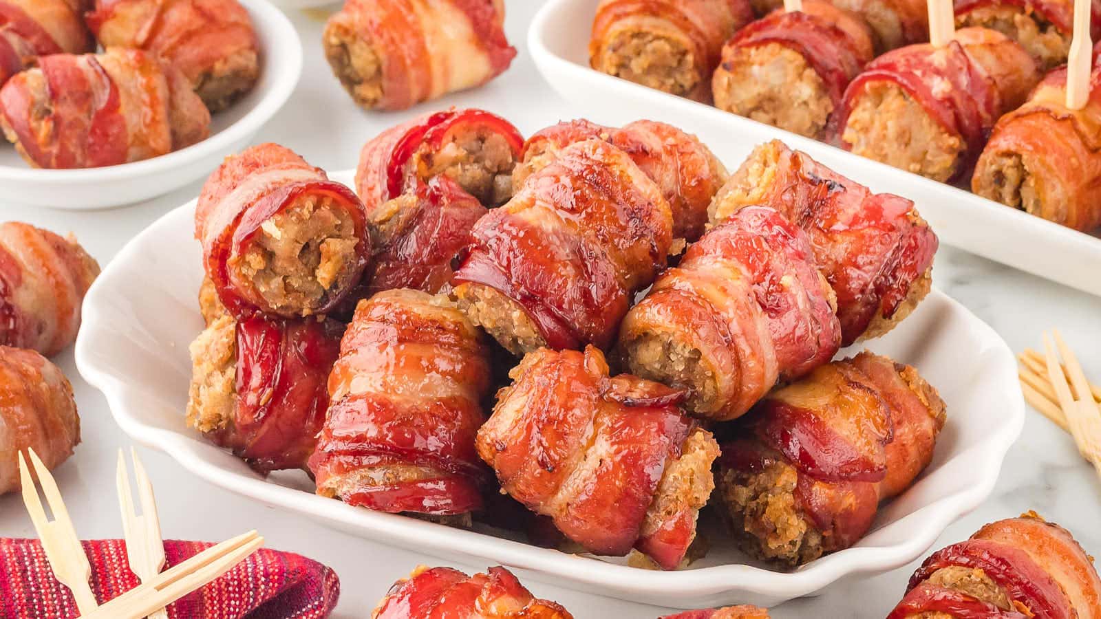 Platter of bacon-wrapped meat appetizers arranged on a white dish, with more servings and wooden picks visible in the background.