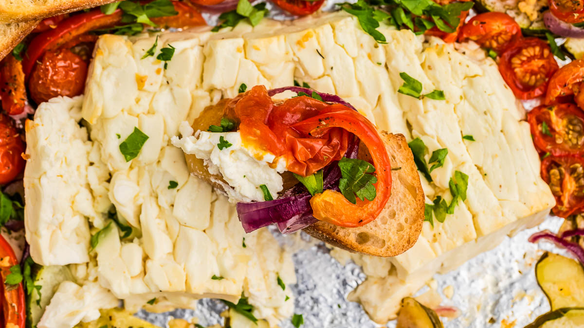 Rectangular block of feta cheese topped with chopped parsley, surrounded by roasted tomatoes and vegetables, with a slice of bread holding cheese, tomato, and onion.
