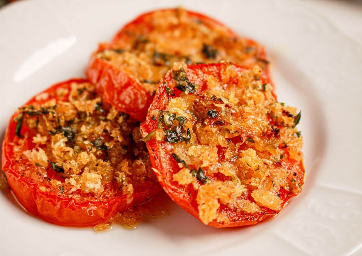 Three slices of baked tomatoes topped with breadcrumbs and herbs are arranged on a white plate.