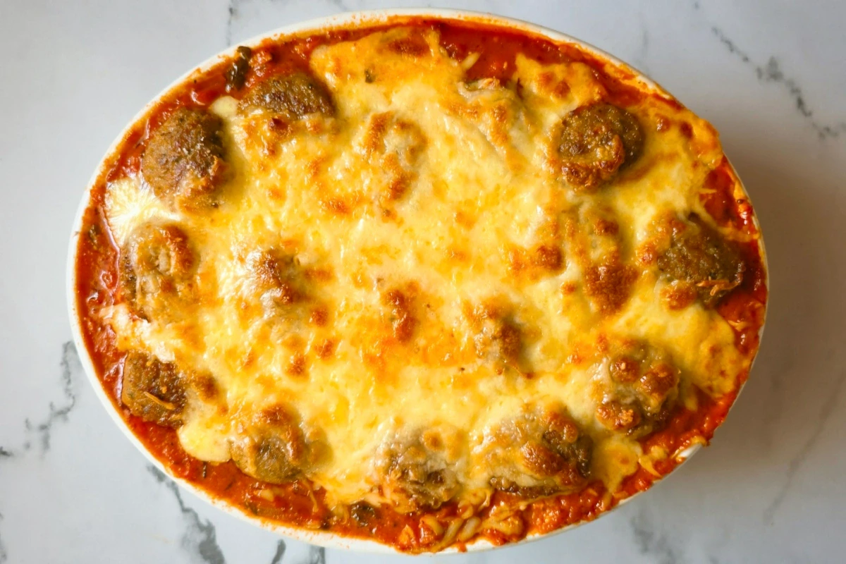 A baked casserole dish filled with meatballs in tomato sauce, topped with melted, golden-brown cheese, set on a marble surface.