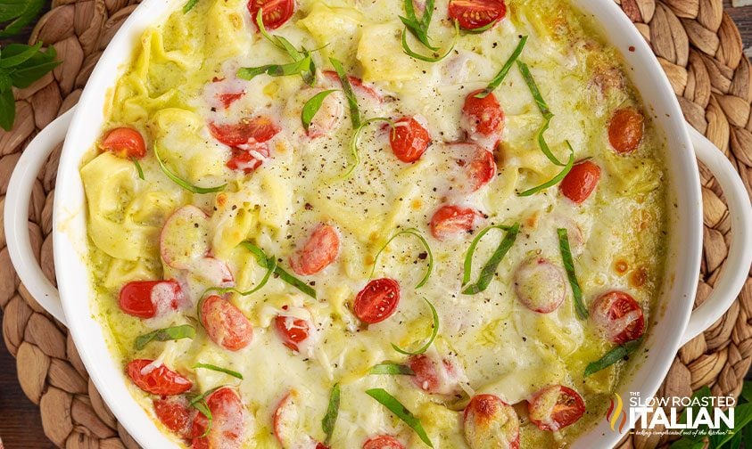 Baked cheese tortellini casserole topped with melted cheese, cherry tomatoes, and sliced basil in a round white dish on a woven mat.