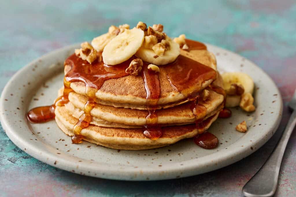 A stack of pancakes topped with banana slices, walnuts, and syrup on a plate, with a fork visible on the side.