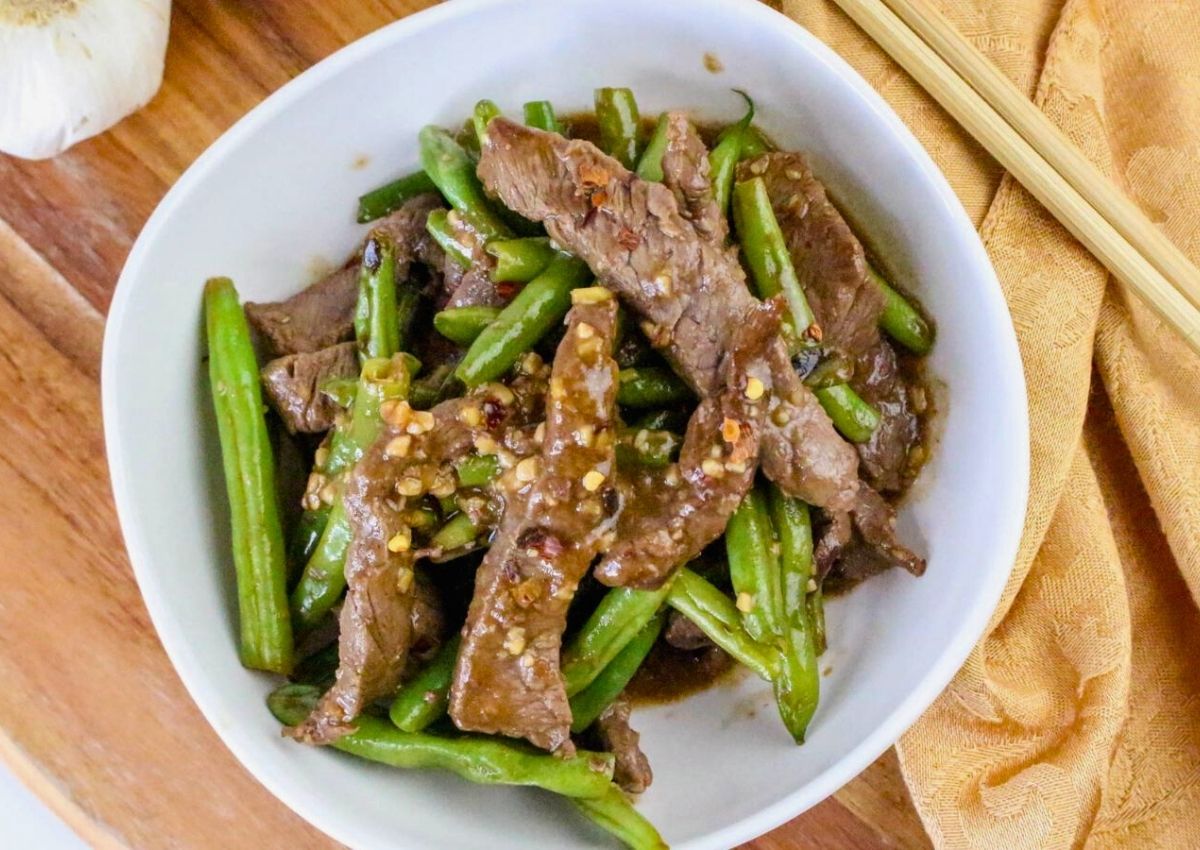 A white bowl filled with stir-fried beef strips and green beans in a savory sauce, placed on a wooden surface with chopsticks nearby.