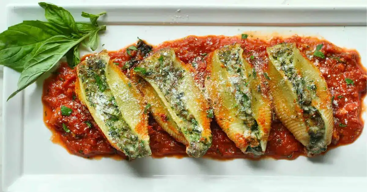 Four stuffed pasta shells with green filling are arranged on a bed of tomato sauce, sprinkled with grated cheese, and garnished with fresh basil on a white rectangular plate.