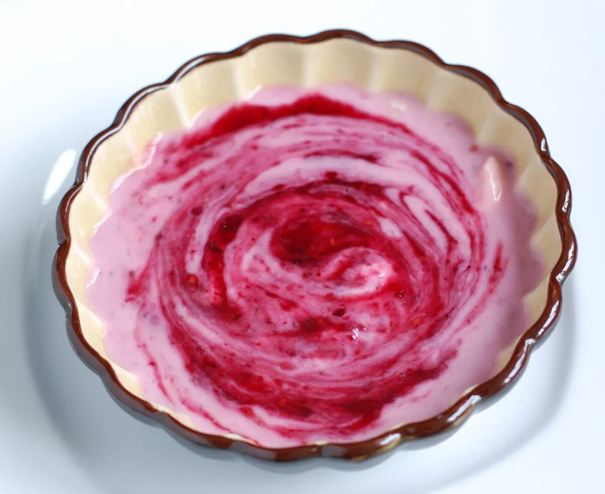 A round beige bowl containing pink yogurt swirled with red berry sauce, placed on a white surface.