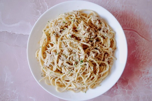 A white plate of spaghetti topped with shredded fish and herbs, placed on a pink textured surface.