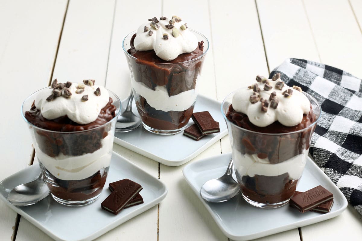 Three glass cups filled with layered chocolate pudding and whipped cream, each topped with whipped cream and chocolate shavings, are served on small plates with spoons and chocolate pieces.
