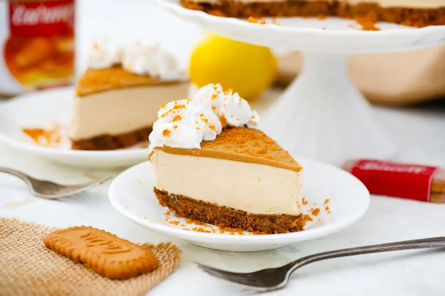 A slice of caramel cheesecake topped with whipped cream and crumbled cookies on a white plate, with a fork and a cookie beside it.