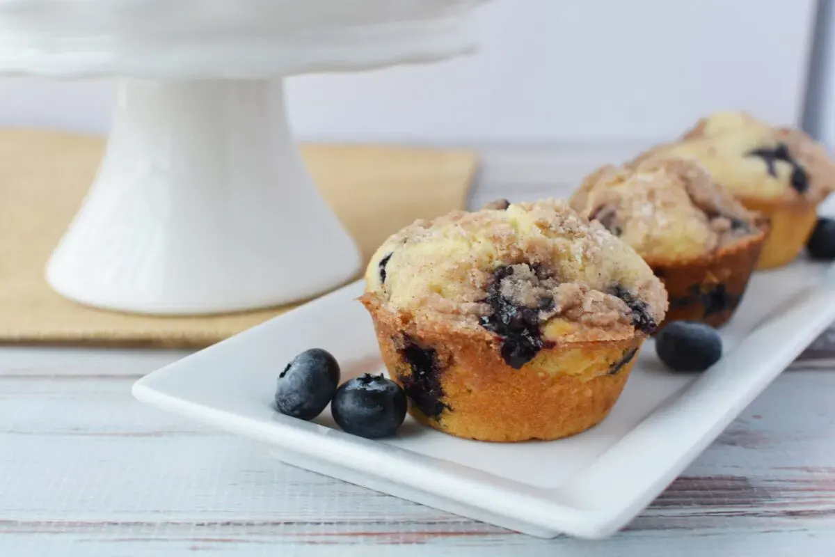 A rectangular white plate holds two blueberry muffins topped with crumb streusel, with a few loose blueberries beside them. A cake stand is in the background.