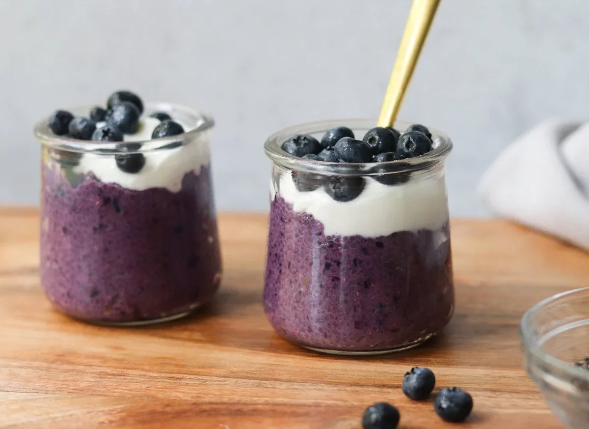 Two glass jars filled with a purple chia pudding, topped with white yogurt and fresh blueberries, are placed on a wooden surface with a spoon in one jar.