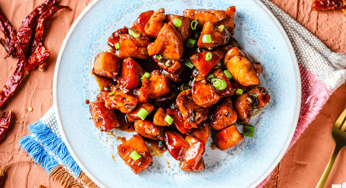 A plate of diced chicken in a dark glaze, garnished with chopped green onions, on a light blue plate atop a woven mat with fringe.