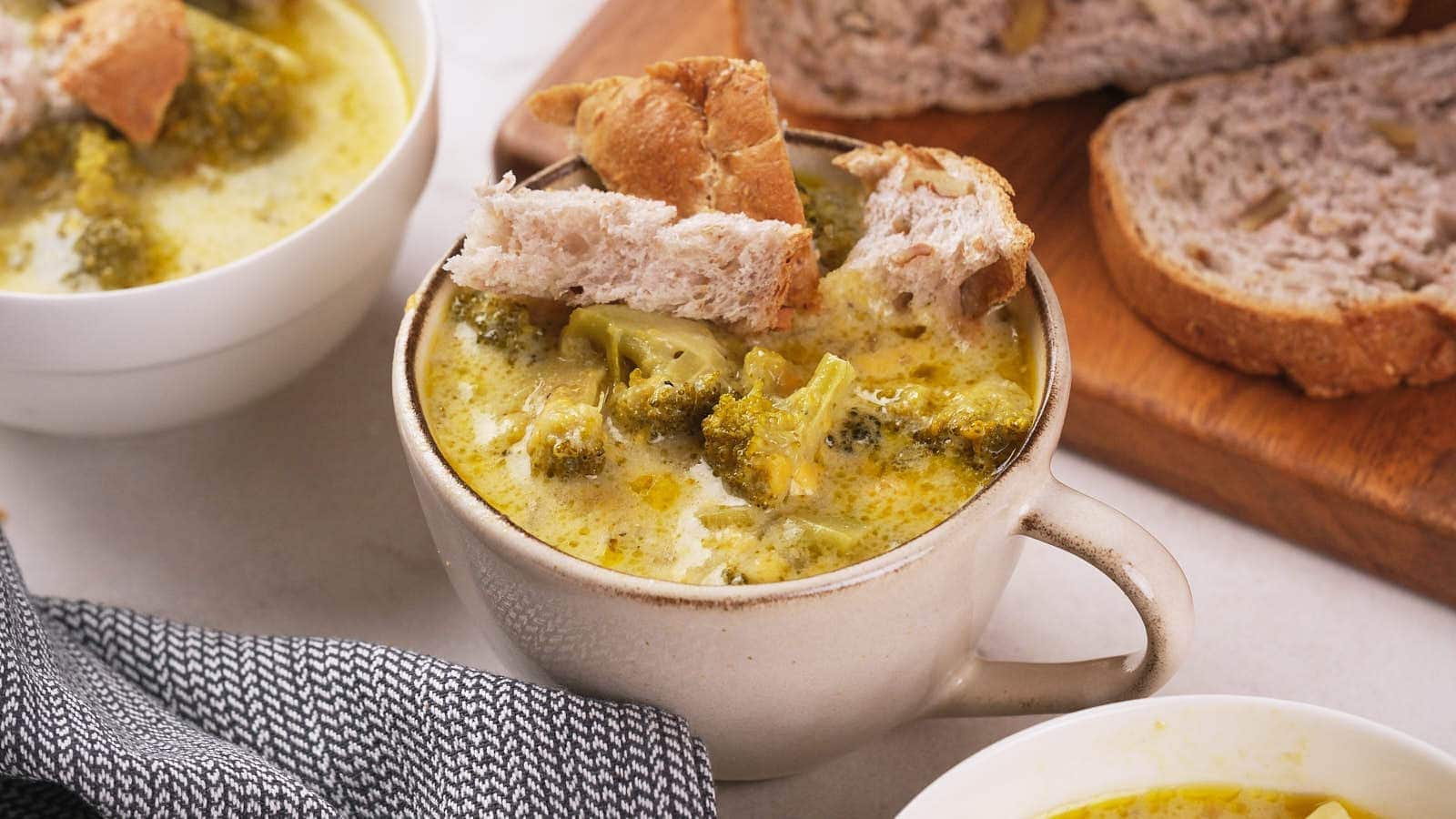 A ceramic mug filled with broccoli soup topped with pieces of bread, with slices of bread and a bowl of soup in the background.
