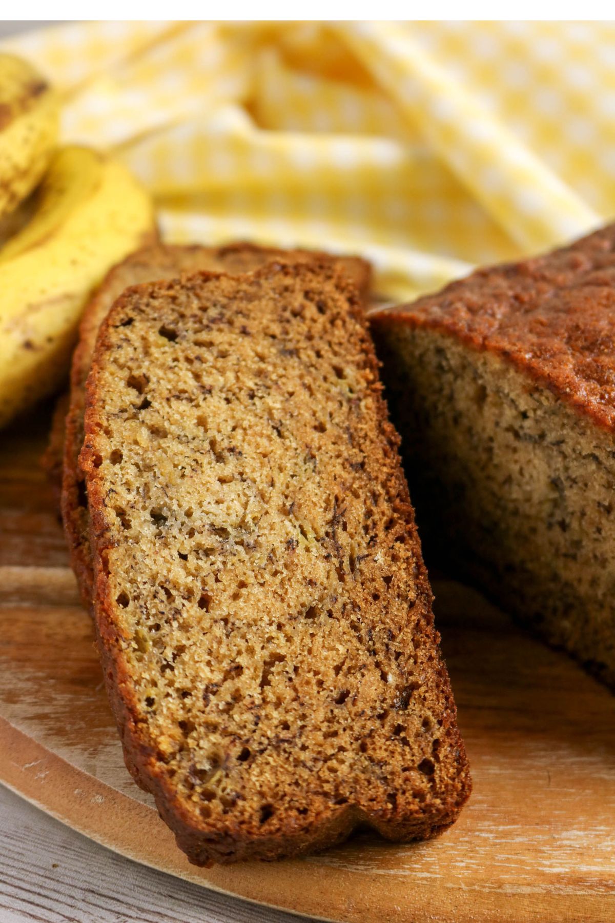 A close-up of a sliced loaf of banana bread on a wooden board, with ripe bananas and a yellow checkered cloth in the background.