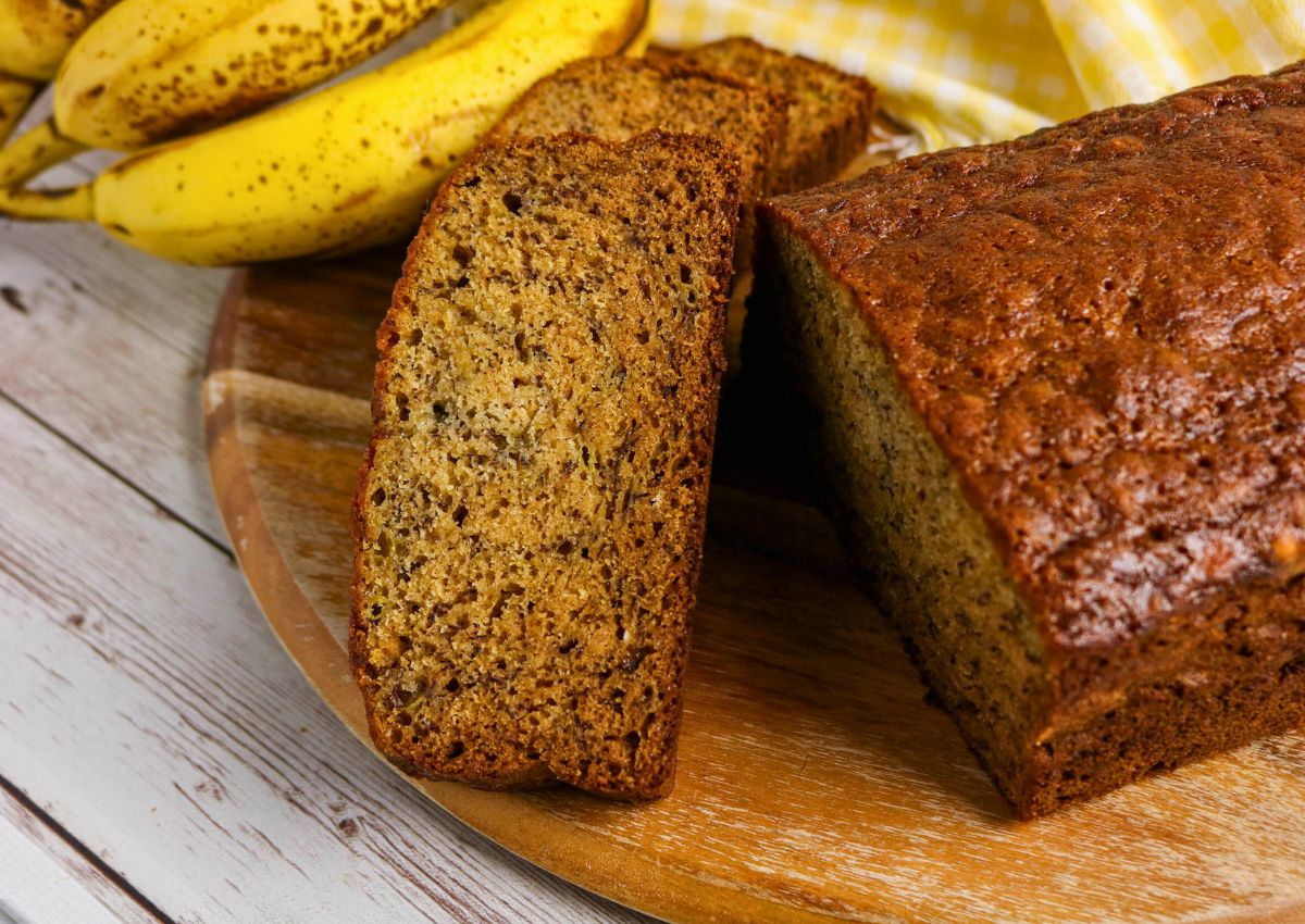 A loaf of banana bread with two slices cut, placed on a wooden board next to ripe bananas and a yellow checkered cloth.