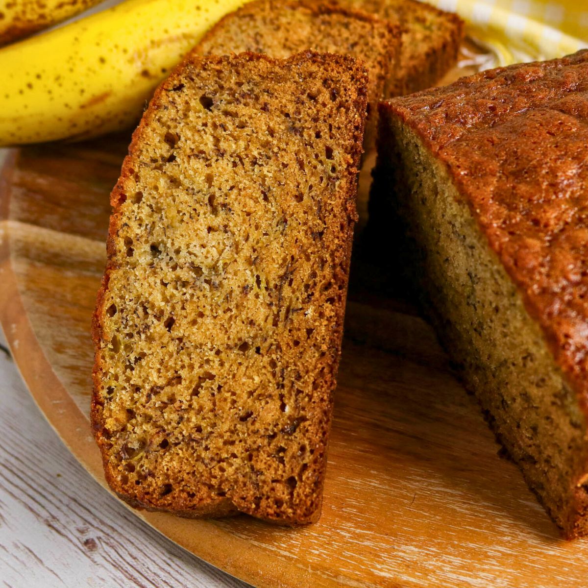 A close-up of a sliced loaf of banana bread on a wooden board with a ripe banana in the background.
