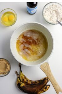 Overhead view of baking ingredients: a pan with melted butter, two eggs in a bowl, flour, brown sugar, vanilla extract, and three ripe bananas on a white surface.