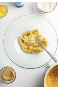 A glass bowl with mashed bananas and a fork, surrounded by ingredients including an egg, brown sugar, flour, and a melted mixture.