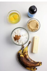Top-down view of baking ingredients: two eggs, vanilla extract, brown sugar, flour with baking soda and cinnamon, a stick of butter, and three ripe bananas on a white surface.