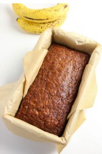 A freshly baked loaf of banana bread in a parchment-lined pan, with two ripe bananas placed behind it on a white surface.