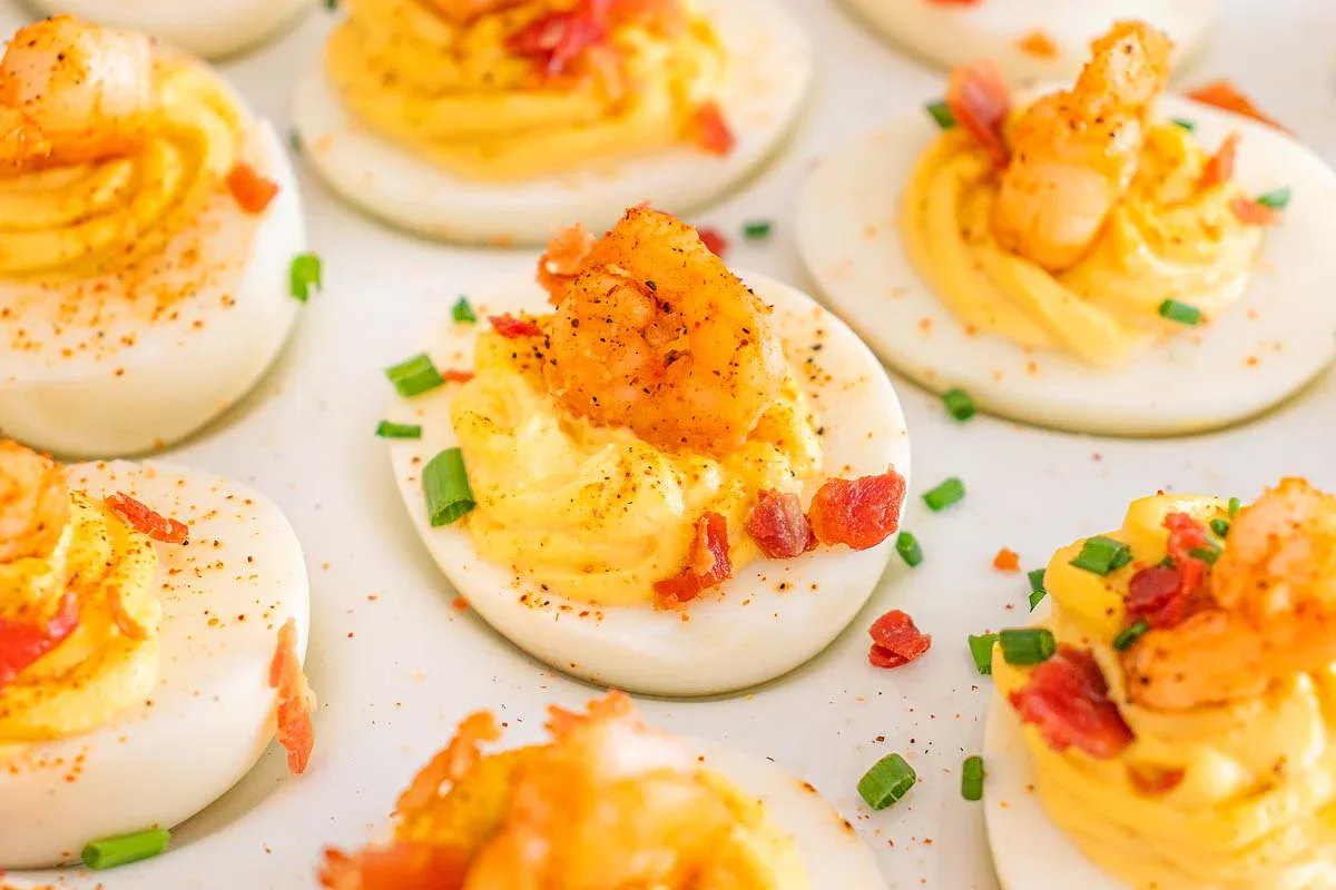 Deviled eggs topped with shrimp, bacon bits, chopped chives, and paprika, arranged on a white surface.
