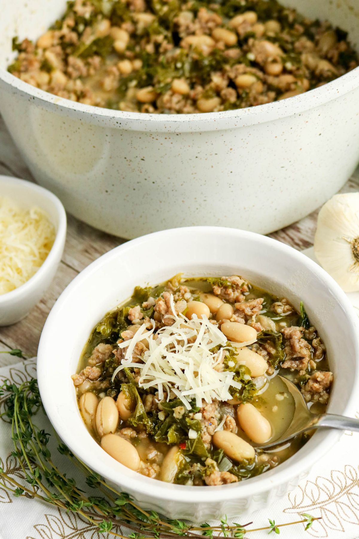 A bowl of white bean and sausage soup with kale, topped with shredded cheese, sits on a table beside a pot of soup, a bowl of cheese, garlic, and fresh thyme.