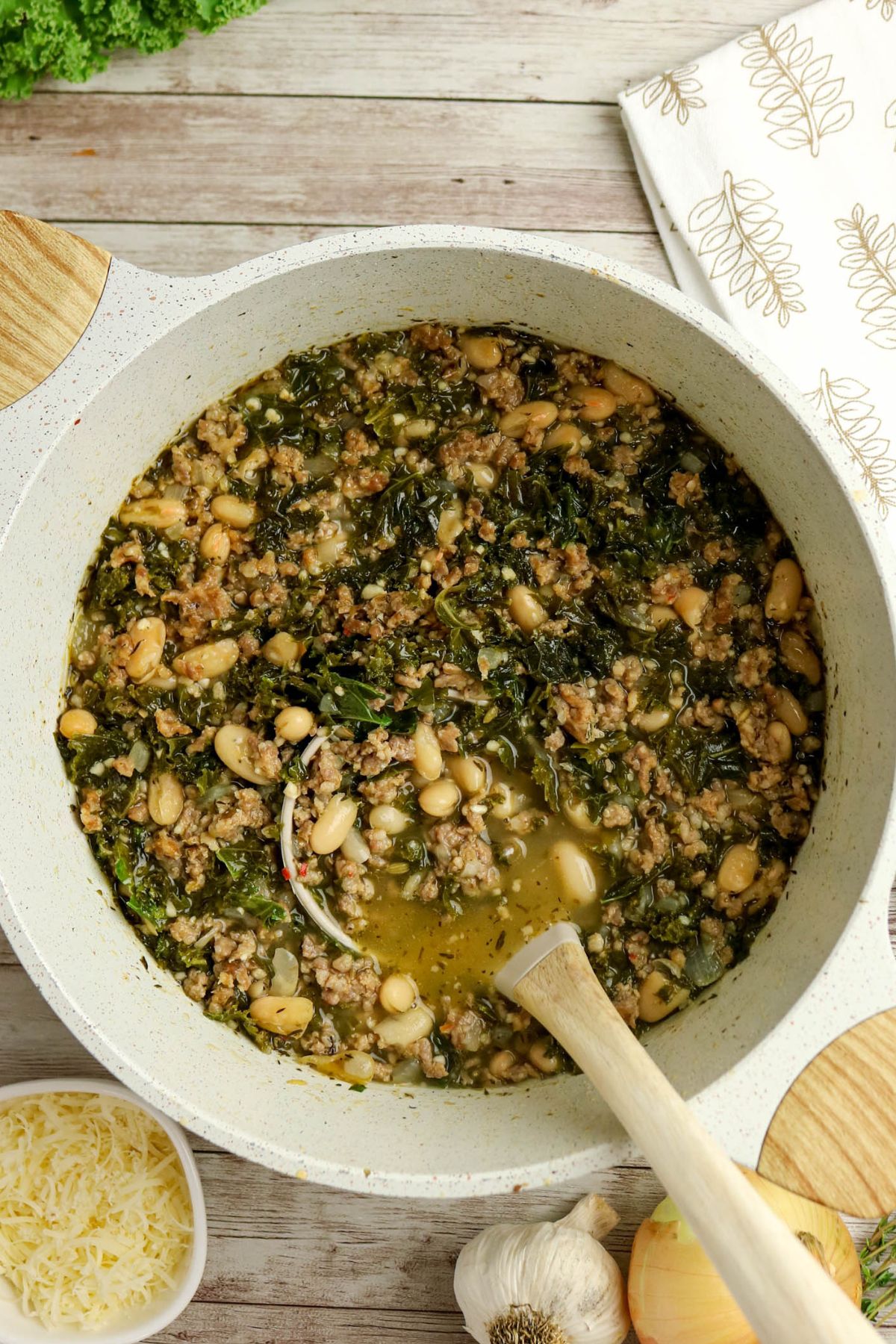 A pot of soup with ground meat, white beans, and kale, alongside grated cheese, garlic, and an onion on a wooden surface.